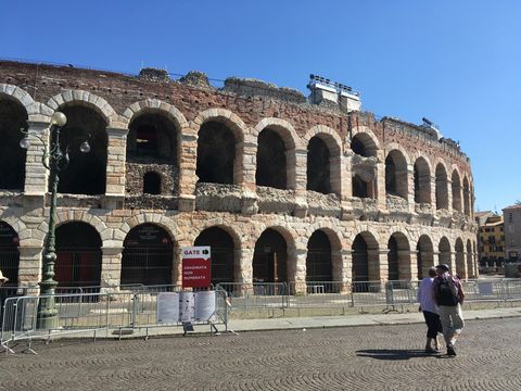 Vista parcial de uma arena antiga, localizada na cidade de Verona. Céu azul sem nuvens. Um casal caminha na rua de pedra logo em frente.