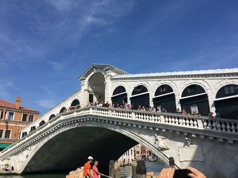 Enorme ponte branca com apenas um arco embaixo. Na parte de cima, muitas pessoas caminhando. Há toda uma construção central ao longo da ponte, com portas pretas espaçadas. No topo, ao centro, há uma área com o telhado mais alto. Céu azul com nuvens brancas.