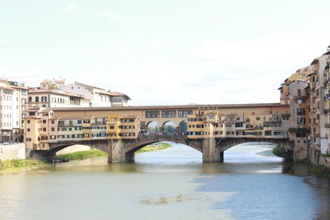 Ponte de três arcos sobre um rio. Em cima da borda da ponte, vê-se uma série de pequenas casinhas, com janelas igualmente pequenas. Em cada ponta da ponte, vários prédios.