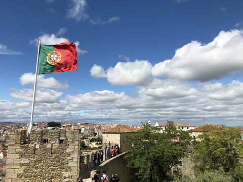 Em primeiro plano, uma torre com a bandeira de Portugal tremulando. Logo atrás, uma muralha ladeada por algumas árvores. Ao fundo, um céu azul cheio de nuvens e o telhado de muitas casas.