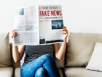 Uma pessoa vestida com blusa listrada em preto e branco e calça jeans está sentada em um sofá cor de creme. Ela lê jornal, que está em frente ao seu rosto. Na manchete, em letras maiúsculas se lê "THE DAILY" (em preto) e "FAKE NEWS" (em vermelho).