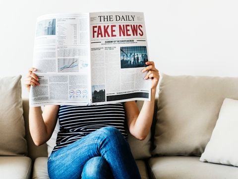 Uma pessoa vestida com blusa listrada em preto e branco e calça jeans está sentada em um sofá cor de creme. Ela lê jornal, que está em frente ao seu rosto. Na manchete, em letras maiúsculas se lê "THE DAILY" (em preto) e "FAKE NEWS" (em vermelho).