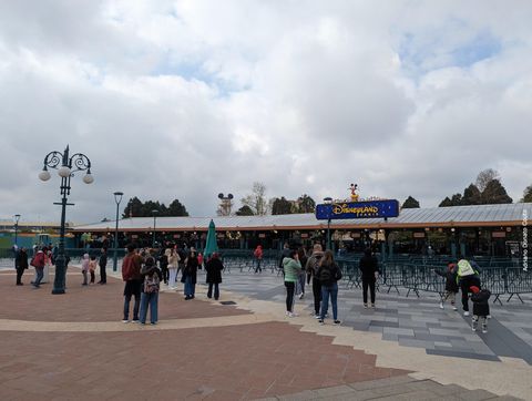 Uma barreira com gradis verdes e catracas, com uma placa azul com os dizeres 'Disneyland Paris' no topo. Algumas pessoas aguardam.
