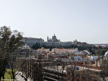 Sob um céu azul claro, vê-se ao fundo um palácio antigo e uma catedral azul claro. Em primeiro plano, várias construções e árvores sem folhas.