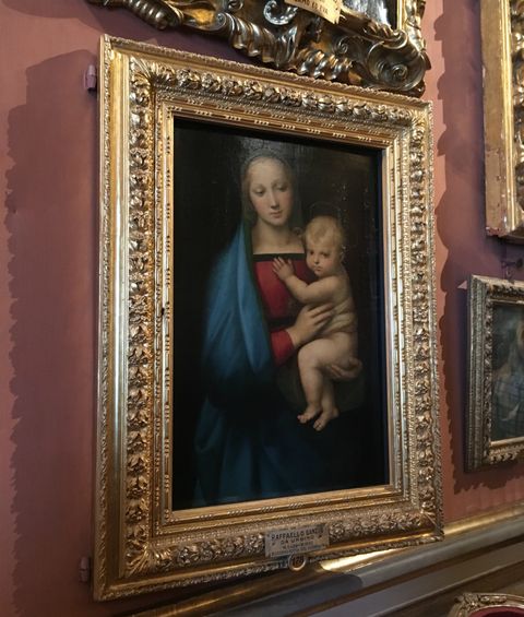 Pendurada em uma parede vermelha, uma pintura com moldura dourada. Na arte, está retratada Nossa Senhora com vestimenta vermelha e manto azul escuro, segurando ao colo o menino Jesus, retrato sem roupas e ainda bebê. A mãe tem um olhar introspectivo, reforçado pelos poucos tons claros da pintura.