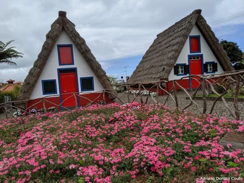 "Duas casas triangulares, com telhado coberto de uma espécie de palha, paredes brancas e portas e janelas vermelhas. Em frente a elas, um jardim de flores rosas."
