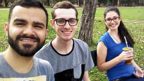 Dois rapazes e uma moça sentados em um banco, rodeados por uma área de gramado. Há parte de um tronco de árvore ao fundo.