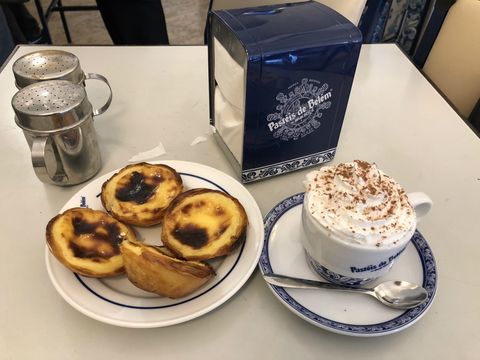 Sobre uma mesa, um porta-guardanapo azul com "Pastéis de Belém estampado em branco. Do lado esquerdo, dois recipientes prateados com açúcar; Em primeiro plano, quatro unidades de pastel de Belém em um prato branco; à direita, uma xícara com creme no topo. Pastéis de Belém têm um formato similar a uma torta, com a cobertura de um creme amarelado com marcas mais escuras do creme tostado.