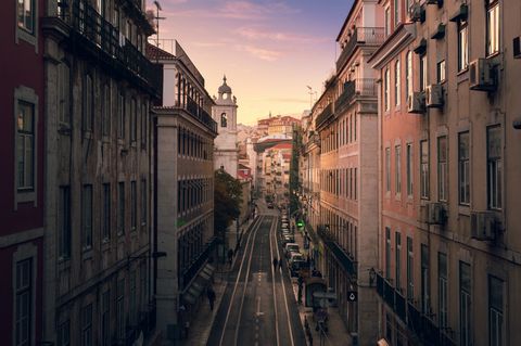 Uma longa rua, ladeada por prédios antigos de vários andares. O céu está amarelado, com um tom que lembra o começo ou o fim do dia.