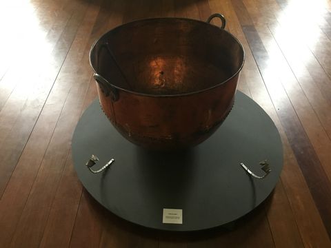 Tacho de cobre sobre círculo cinza escuro. O conjunto está sobre um piso de madeira.