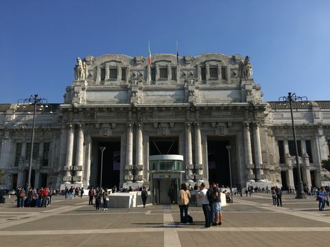Em uma grande área aberta, várias pessoas espalhadas. Ao fundo, um prédio com três portas gigantes abertas, ornamentado com longas colunas de pedra e algumas estátuas no topo. Céu limpo sem nuvens.