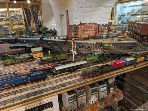 'Em uma sala, vemos um grande modelo em miniatura com trilhos, trens e prédios antigos.'